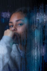 Foto de una mujer que se ve a través de un cristal lleno de gotas de agua como si estuviera lloviendo, la chica tiene la mano apoyada en la cara y con semblante pensativo. Como tratar la ansiedad, como tratar la depresión, tratar el estrés, alimentación sana, relación del nervio vago con la ansiedad, como cuidar el nervio vago, técnicas de relajación. 